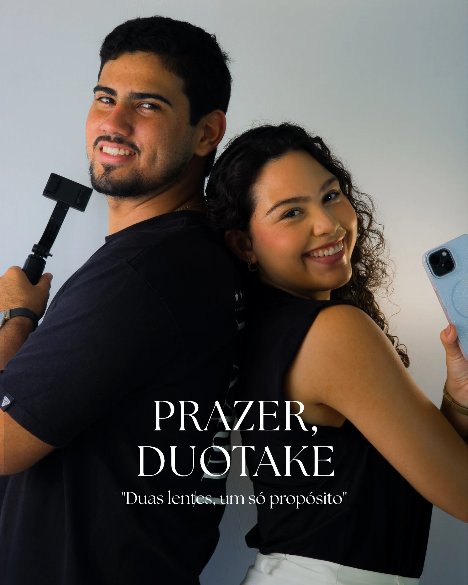 O casal da DUOTAKE, de costas um para o outro, ele com o bastão de celular e ela com o celular na mão